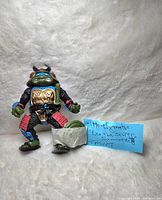 Samurai Leonardo action figure posed with sword and shield, placed on a white fur background with a handwritten note describing the item.