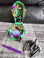 Opened Joker's Head playset showing interior with two figurines, a bat vehicle, and a purple weapon on gray carpet with black background.