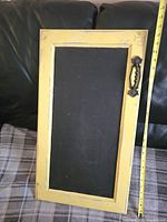 Full view of the vintage cupboard door chalkboard showing the yellow distressed wooden frame and black chalkboard center with ornate black handle on the right side placed vertically on couch.