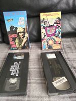 Photo of two vintage VHS movies "Sands of Iwo Jima" and "UHF" with their original sleeves and tapes laid out on a fabric surface.