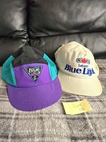 Photo of two vintage Labatt beer promotional hats, one Labatt Blue and one Labatt Blue Light, placed side by side on a fabric surface with black leather couch background.