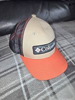 Side view of the youth Columbia hat showing tan front, black mesh sides, orange brim and embroidered logo patch.