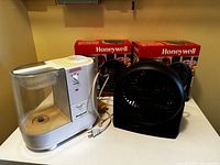 Two black Honeywell table fans in original boxes alongside white Honeywell humidifier on white surface.