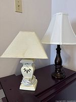 Two table lamps shown side by side on dark wood surface, ceramic lamp cream-colored with trapezoidal shade and burnished copper finish lamp with bell shade