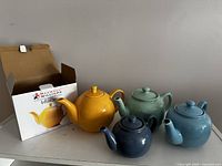 Four colorful ceramic teapots in mustard yellow, sage green, sky blue, and navy blue, with original packaging for the mustard yellow one.