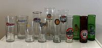 Front row of 14 different branded beer glasses varying height and color including Coors Light, Hoegaarden, Miller Lite, Erdinger, Amstel, and others.