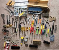 Top down view of assorted hand tools laid out on carpet including saws, screwdrivers, wrenches, pliers, brushes, and small hardware compartment boxes with nails and screws.