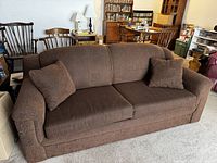 Front view of brown plush sofa bed with two matching cushions on a beige carpet within a furnished room.