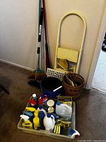Wide view of cleaning supplies in a plastic tote with various cleaning liquids, brushes, and sponges. Cleaning tools and wastebaskets arranged behind tote.