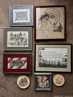 Seven distinct framed needlework cross stitch pieces laid out together on carpet, showing various designs including a dreamcatcher, farm animals, a red house, text, cat, and floral patterns.