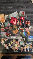 Lot of assorted fridge magnets featuring Coronation Street characters and various European country souvenirs laid out on dark wooden floor.
