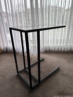 Angled side view showing black metal frame with tempered glass top, placed on carpet in front of sheer curtains.