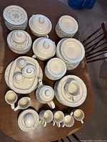 Image of a large collection of Cordella stoneware dishware laid out on a wooden table including plates, cups, saucers, lidded serving bowls, teapot, and various serving pieces.