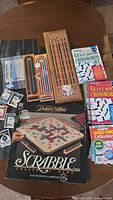 Lot displayed on a wooden table featuring the Scrabble Deluxe Edition box, three cribbage boards, and three puzzle books.