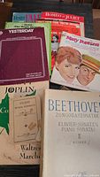 Top view of assorted vintage sheet music with colorful and monochrome covers including Mary Poppins, Romeo & Juliet, Beethoven, and Yesterday titles