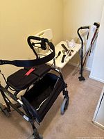 Image showing Nexus collapsible walker with black seat cushion and storage basket, two walking canes, accessories on a table, and over edge bath chair in corner.