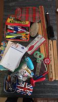 Top-down photo showing various office, sewing, and stationery items including wooden yardstick and rulers, brushes, pens, sewing kit, tape dispensers, and keys.