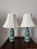 Front view of two teal ceramic table lamps with white bell-shaped shades, showing the lamp shape and color.
