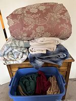 Three comforters and three cotton blankets stacked on a wooden surface above a blue plastic storage tub filled with folded sheet sets in solid colors.