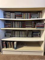 Front view of white wooden shelf with CDs arranged on three shelves plus bottom shelf with fewer CDs