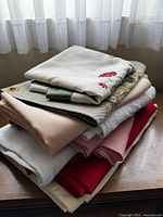 Stack of assorted tablecloths and runners showing various fabrics, colors, and designs including floral embroidery and lace.