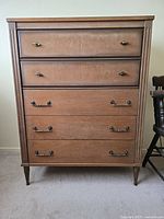 Front view of vintage tall wood dresser, showing five drawers with metal handles, tapered legs, and natural wood finish.