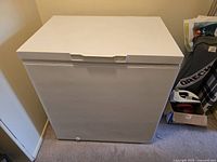 Closed white Whirlpool chest freezer with smooth finish and integrated lid handle, placed against a wall with surrounding household items.