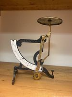 Side angle of the antique balance scale showing brass and cast iron components, engraved details, white measurement dial, and circular weighing platform.