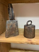 Photo of cast iron cow bell with worn leather strap and iron weight on wood shelf.