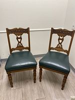 Pair of antique wooden accent chairs with carved backs and dark green leather upholstered seats, standing side by side.