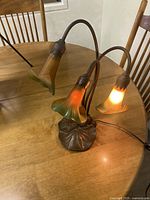 Overall view of the Art Nouveau table lamp showing three lily-shaped glass shades with warm orange and green coloration, lit on a wooden table.