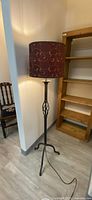 Overall view of the floor lamp showing the burgundy fabric shade with gold scrollwork and black metal tripod base standing near a wall and wooden shelving unit.