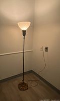 Full view of the lit floor lamp in corner with power cord extended, showing height and glass shade.