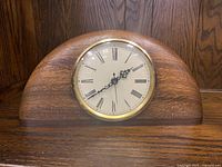 Front view showing wooden half-moon shape mantel clock with Roman numerals and brass bezel glass cover.