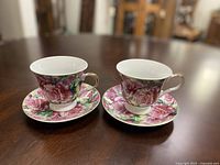 Two porcelain teacups each decorated with a pink rose floral design on a wooden table.
