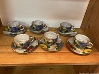 Six floral pottery teacups and matching saucers displayed on a wooden shelf, showing colorful flower designs on each piece.