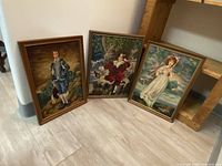 Three framed cross-stitch portraits arranged leaning against a wooden shelf on a tiled floor, colors and details visible, showing vintage and classical styling.