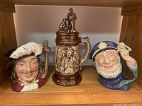 Three ceramic mugs on wooden shelf showing full set and details of designs: historical figure, lidded beer stein, nautical sailor mug