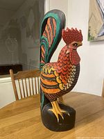 Front-left view of the wooden rooster sculpture showing detailed painted feathers and red comb with white dots.