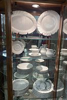Photo of the full set including plates, bowls, teacups, saucers, and platter inside a glass cabinet, showing edge detailing and embossed pattern.