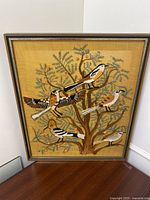 Front view of embroidered wall art with birds and tree design on mustard yellow fabric in wooden frame.