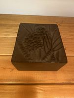 Closed square metal box showing raised pine cone pattern on lid