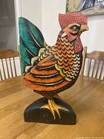 Front angled view showing multicolor painted wooden rooster sculpture with intricate feather pattern and red comb on black base.