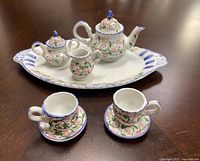 Miniature porcelain tea set displayed on a wooden surface, showing the teapot, creamer, sugar bowl with lid, two cups on saucers, and serving platter. Floral design with pink, green, and blue accents.
