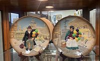 Two Royal Doulton character plates displayed on a shelf, showing the two figures of old balloon sellers with raised 3D colorful designs against a cityscape background.