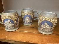 Set of three ceramic beer steins showing beige and blue relief decoration and sturdy handles