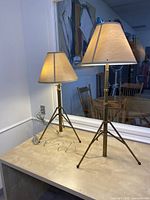 Two brass finish adjustable tripod base table lamps with beige fabric shades placed on a light wooden surface in front of a windowed wall.