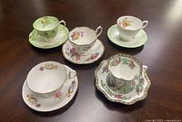 Five bone china teacups with unique delicate floral designs and matching saucers arranged on a wooden surface.