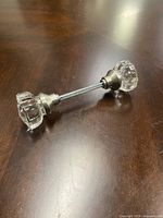 Two clear faceted glass door knobs with silver metal bases connected by a threaded metal spindle, shown on wooden surface from angles to display shape and details.