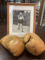 Pair of vintage tan leather boxing gloves placed in front of a framed black-and-white photo of a boxer in pose, on a table indoors